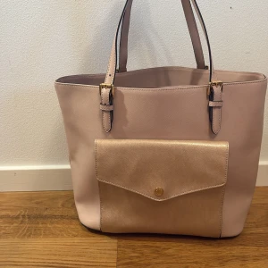 Beige axelväska från Michael Kors - Stilren ljus råsa Michael Kors i skinn med justerbara axelband och guldfärgade detaljer. Väskan har en stor ficka framtill med tryckknapp och rymligt innerfack för dator och med flera mindre fack och dragkedja. Perfekt för dig som vill ha plats för allt du behöver.