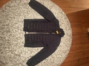 Svart dunjacka från Parajumpers - Säljer en svart dunjacka från Parajumpers med gul detalj vid halsen och patch på ärmen. Jackan har quiltad design, dragkedja framtill och långärmade ärmar. Insidan är ljusgrå och jackan är lätt och smidig, perfekt för kyliga dagar.