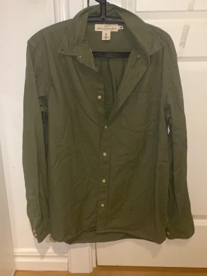 Olivgrön skjorta från L.O.G.G. i bomull - Säljer en olivgrön skjorta från L.O.G.G. i storlek L. Skjortan är tillverkad i mjuk bomull, har klassisk krage, bröstficka och knappar framtill. Perfekt för en avslappnad och stilren look. Passar dig som gillar enkel och tidlös stil.