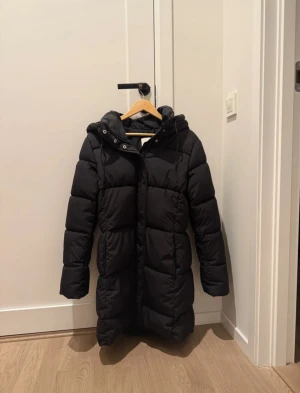 Svart lång pufferjacka med huva - Säljer en svart pufferjacka med huva och snörning. Jackan är lång och med dragkedja framtill. Perfekt för kalla dagar! Den går ner till låren, köpte den av en tjej på Vinted och den var tyvärr för stor! 