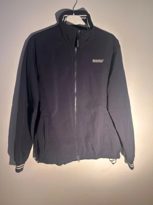 Mörkblå  vindjacka från Woolrich - Svart vindjacka från Woolrich med hel dragkedja och ståkrage. Jackan har vita ränder på mudden vid ärmslut och insidan av kragen, samt diskret logga på bröstet. Tillverkad i lätt polyester, perfekt för blåsiga dagar. Snygg och enkel design med normal passform.