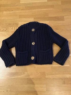 Mörkblå stickad kofta med stora knappar - Säljer en chunky mörkblå stickad kofta med tre stora silvriga knappar framtill och två praktiska fickor. Koftan har långärmad passform och grovstickad struktur som ger en cool och mysig vibe. Perfekt för att hålla värmen och se snygg ut.