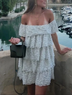 Vit prickig offshoulder klänning - Superfin vit kortklänning med svarta prickar och flera lager volanger. Offshoulder-modell som ger en chill och trendig vibe. Perfekt för varma dagar och har en luftig passform. Materialet känns lätt och bekvämt mot huden. OBS lappen är bortklipp 