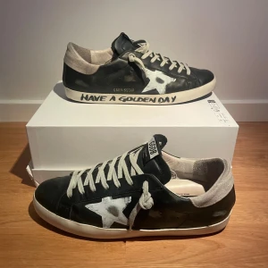 Golden Goose svarta sneakers (sällsynta) - Svarta Golden Goose sneakers i skinn med vit stjärndetalj på sidan, grå mockadetaljer vid hälen och vita snören. Sulan har texten 'Have a Golden Day' 