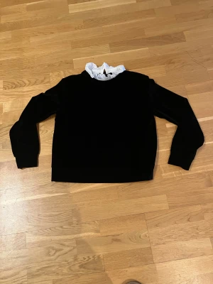 Svart sammetströja med vit volang - Snygg svart långärmad tröja i mjuk sammet med en vit volangkrage som ger en cool kontrast. Perfekt för dig som gillar att sticka ut med detaljer. Tröjan har en klassisk passform och känns både trendig och bekväm.