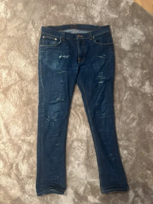 Blå jeans från Nudie Jeans med slitningar - Säljer ett par blå jeans från Nudie Jeans med coola slitningar och kontrastsömmar. Klassisk femficksmodell med broderade bakfickor och läderpatch i midjan. Jeansen har rak passform och är tillverkade i robust denim. Pris inte hugget i sten!