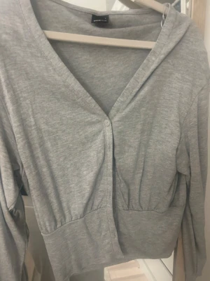 Grå kofta från Gina Tricot - Ljusgrå kofta från Gina Tricot i mjuk bomullsblandning. Modellen har v-ringning, långa ärmar och knäppning framtill med tre knappar. Perfekt att slänga över en topp för en chill look.