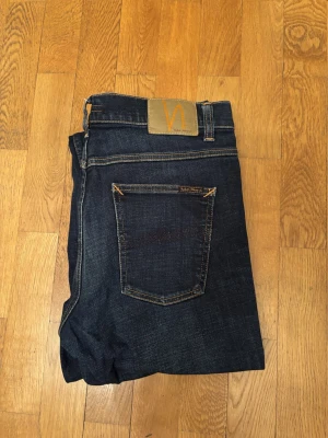 Mörkblå jeans från Nudie Jeans - Snygga mörkblå jeans från Nudie Jeans med klassisk femficksdesign och kontrastsömmar. Modellen har slim fit passform och är tillverkad i slitstarkt denimtyg av bomull. Läderpatch med Nudie-logga baktill i midjan. Perfekta för dig som gillar stilrena och tidlösa jeans. Modellen heter Lean Dean. Hör av er vid minsta fundering!
