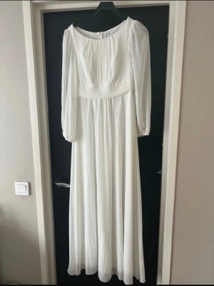 Vit långklänning  - Säljer en elegant vit långklänning  i storlek M. Klänningen har långärmade puffärmar, rund halsringning och smockad midja som ger en snygg siluett. Materialet är mjukt och faller fint, perfekt för dig som gillar klassisk och stilren look.
