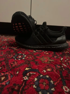 Svarta Adidas Ultraboost sneakers - Säljer ett par helsvarta Adidas Ultraboost sneakers med stickad ovandel och klassiska tre ränder på sidan. Skorna har snörning, platt sula och en riktigt bekväm Boost-mellansula. Perfekta för dig som gillar stilrena och sportiga skor.