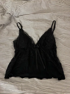 Svart spetstopp - Säljer en svart linnetopp med tunna axelband och v-ringad spets framtill. Toppen har en söt rosett i mitten och detaljerade spetskanter längs sidorna. Materialet är mjukt och glansigt, troligen polyester eller satin, och toppen är ärmlös med en lätt croppad passform.