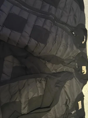 Svart dunväst från Moncler - Snygg svart dunväst från Moncler med quiltad design och ribbade detaljer på fickorna. Västen har dragkedja framtill och en stilren, sportig look som passar perfekt till lager-på-lager. Materialet är dun och polyester för extra värme och komfort.