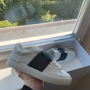 Valentino Garavani vita sneakers - Snygga vita Valentino Garavani sneakers i skinn med bred svart detalj över sidan och coola nitar bak på hälen. Klassisk rund tå, platt sula och vita snören. Perfekta för dig som vill ha en stilren men ändå unik look.