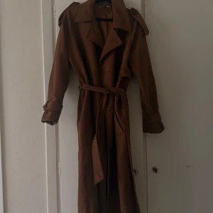 Brun trenchcoat med bälte - Snygg brun trenchcoat med klassisk krage, axelklaffar och bälte i midjan. Jackan har långa ärmar med knappar vid mudden och är tillverkad i ett mjukt mockaliknande material. Perfekt för dig som vill ha en stilren och tidlös look.