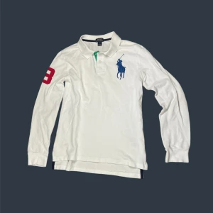 Vit långärmad pikétröja Polo Ralph Lauren - Vit långärmad pikétröja från Polo Ralph Lauren med blå broderad logga på bröstet och röd siffra 3 på ärmen. Klassisk krage med knappar och grönt tygdetalj vid knappslån. Skön bomullskvalitet och normal passform. Står L men passar S