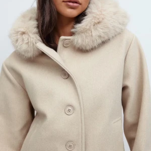 Beige kappa med fluffig krage - Säljer en beige kappa med päls på huvan. Jackan har en rak, loose passform och stängs med stora knappar framtill. Endast använd en gång! Slutsåld i denna storleken🤗