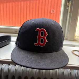 Boston Red Sox keps från New Era - Snygg svart keps från New Era med Boston Red Sox logga broderad i rött och vitt framtill. Klassisk rak skärm och broderad New Era-logga på sidan. Perfekt för dig som gillar sportig streetstyle.