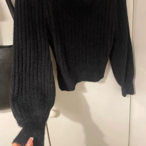 Svart ribbad stickad tröja Minkpink - Mysig svart stickad tröja från Minkpink med ribbat mönster och fyrkantig ringning. Tröjan har långa ärmar med lite puff och sitter snyggt på kroppen. Perfekt för kyliga dagar och enkel att styla med jeans eller kjol.