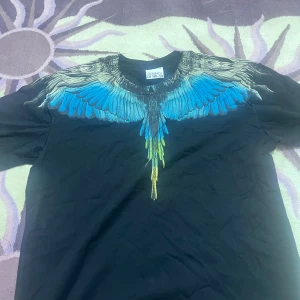 Svart t-shirt Marcelo Burlon med vingar - Svart t-shirt från Marcelo Burlon County of Milan med ikoniskt vingtryck i blått, svart och gult över axlar och bröst. Klassisk passform och rund hals. Perfekt statement-plagg för dig som vill sticka ut.