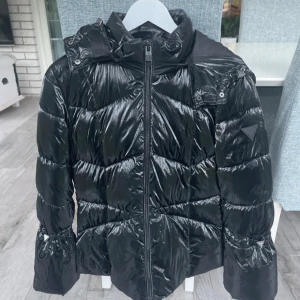 Svart pufferjacka från Guess - Snygg svart pufferjacka från Guess med glansig finish och klassisk logga på ärmen. Jackan har huva, dragkedja framtill och quiltad design som ger en cool look. Perfekt för kalla dagar när du vill hålla stilen.