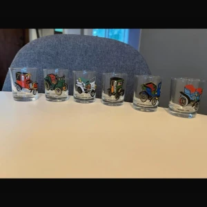 Set med 6 vintage snapsglas med klassiska bilar - Unikt set med 6 snapsglas, dekorerade med färgglada motiv av klassiska bilar från 1899-1907. Glasen är i fint skick utan synliga skador eller slitage. Perfekt för samlare eller som present till bilentusiasten.