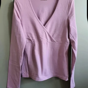 Ljuslila omlottopp från Vero Moda - Supersöt ljuslila omlottopp från Vero Moda med lång ärm och v-ringning. Tillverkad i mjuk bomullsmix som känns skön mot huden. Perfekt att styla med jeans eller kjol för en chill look.