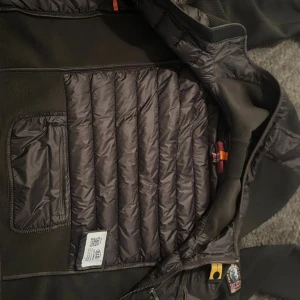 Mörk grön puffer cardigan Parajumper - Säljer en mörk grön puffer cardigan från Parajumpers med quiltade partier och huva. Jackan har dragkedja framtill, diskreta fickor och coola detaljer som orange hängare och patch på ärmen. Perfekt för kyliga dagar när du vill hålla stilen.