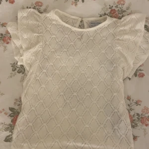 Vit blus från Holly & Whyte - Supersöt vit blus från Holly & Whyte (Lindex) med korta volangärmar och ett diskret rutmönster i tyget. Blusen har rund halsringning och knäppning bak i nacken. Perfekt för dig som gillar romantiska detaljer och en luftig känsla.