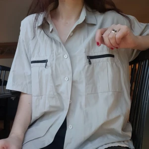 Beige kortärmad skjorta med dragkedjor - Beige skjorta med korta ärmar och avslappnad passform. Två bröstfickor med svarta dragkedjor ger lite utility-vibe. Skjortan har klassisk krage och knäppning framtill. Jag har vanligtvis storlek 36 och denna är som ni ser lös på mig. 💝