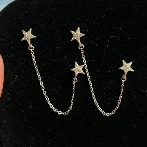 Silvriga örhängen med stjärnor och kedja - Säljer ett par snygga silvriga örhängen med glittriga stjärnor och tunn kedja som kopplar ihop två hål i örat. Perfekt för dig som vill sticka ut lite extra med din stil. Stjärnorna är täckta av små stenar som ger extra bling.