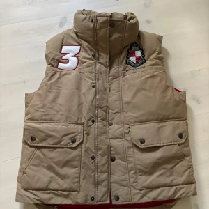 Berkeley Väst - Säljer en beige dunväst från Berkeley med röd insida och broderad logga på bröstet. Västen har hög krage, stora fickor med lock och tryckknappar samt dragkedja framtill. Snygg detalj med stor siffra 3 på axeln och emblem på bröstet.