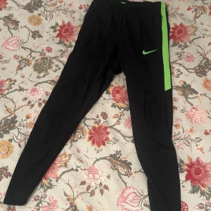 Nike Dri-Fit svarta träningsbyxor S - Svarta Nike Dri-Fit träningsbyxor med grön detalj och logga på benet. Tight passform och elastisk midja. Tillverkade i polyester och elastan för maximal komfort vid träning. Perfekta för dig som vill ha sportiga och snygga träningsbyxor. Byxorna har dragkedja längst ner vid bensluten. Passar även dig som har xs i vanliga fall.