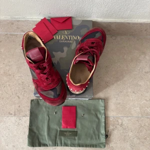 Valentino Rockrunner sneakers röd/camo - Säljer ett par Valentino Rockrunner sneakers i rött och kamouflage med coola nitar runt sulan. Skorna har en mix av mocka och textil, rund tå och svart platt sula. Kommer med originalpåse och tillbehör (extra spikes). Stl 37 Perfekta för dig som vill sticka ut med lyxig streetstyle inför de kalla vädret. 