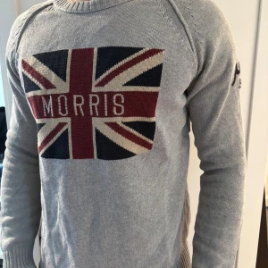 Grå stickad tröja från Morris - Snygg grå stickad tröja från Morris med Union Jack-flagga och stor logga framtill. Tröjan har rund hals, ribbade muddar och svart broderad detalj på ena ärmen. Perfekt för dig som gillar klassisk stil