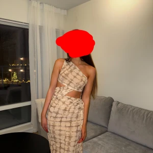 Beige oneshoulder långklänning - Trendig beige långklänning med oneshoulder och cut-out detaljer i midjan. Klänningen har ett unikt batikmönster och omlottkänsla vid höften. Perfekt för dig som vill sticka ut med en cool och modern look.