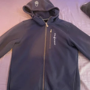 Mörkblå hoodie från Sail Racing - Mörkblå hoodie från Sail Racing med dragkedja framtill och bröstficka med logga. Tröjan har huva med snörning och är tillverkad i ett mjukt, fleeceliknande material. Perfekt för kyliga dagar och snygg till jeans eller träningsbyxor. 