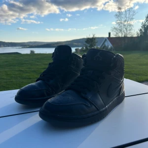 Svarta Nike Air Jordan 1 High - Säljer ett par helsvarta Nike Air Jordan 1 High sneakers i skinn. Klassisk siluett med snörning, rund tå och ikoniska Jordan-loggor på sidan och hälen. Perfekta för dig som gillar streetwear och vill ha en stilren look.