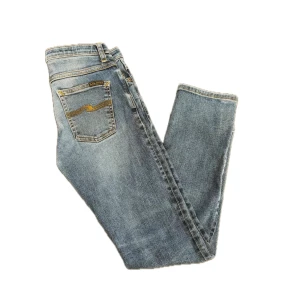 Nudie Jeans Skinny Blå Ripped - Snygga blå skinny jeans från Nudie Jeans med slitningar och hål på knäna. Klassisk femficksmodell med gul brodyr på bakfickorna och normal midja. Perfekt för dig som gillar en avslappnad men trendig look.