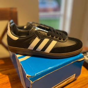 Adidas Samba svart/vit sneakers - Klassiska Adidas Samba sneakers i svart och vitt med de ikoniska tre ränderna på sidan. Ovandel i skinn och mocka, bruna gummisulor och svart snörning. Perfekta för dig som gillar retrostil och streetwear. Snygg detalj med guldfärgad Samba-text på sidan. Priset är inte hugget i sten 
