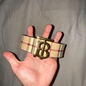 Burberry rutigt bälte med guldspänne - Säljer ett brett Burberry-bälte i klassiskt beige rutigt mönster med inslag av rött och svart. Bältet har ett stort guldigt 'B'-spänne och är gjort i skinn. Perfekt accessoar för att lyfta din outfit med en lyxig touch.