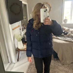 Mörkblå pufferjacka Polo Ralph Lauren XL - Mörkblå pufferjacka från Polo Ralph Lauren i storlek XL. Jackan har huva, dragkedja framtill och två sidofickor med dragkedja. Klassisk Polo-logga broderad på bröstet. Perfekt för kalla dagar och riktigt snygg passform.
