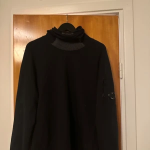 Svart polotröja från C.P. Company - Svart polotröja från C.P. Company med coolt emblem på ärmen och ribbad detalj vid halsen. Tröjan har lång ärm och en hög krage som ger en snygg streetwear-vibe. Perfekt för dig som gillar stilrena och moderna plagg.