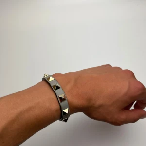 Grått armband med nitar - Snyggt grått armband i skinn med silvriga pyramidformade nitar runt om. Justerbart spänne i metall för perfekt passform. Perfekt accessoar för att ge din outfit en edgy vibe.