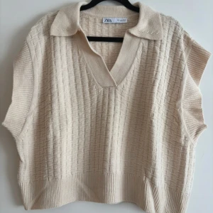 Beige stickad topp från Zara - Beige stickad väst topp från Zara med krage och v-ringning. Toppen har ett diskret rutmönster i strukturen och är ärmlös med breda ribbade kanter. Perfekt för lager-på-lager och ger en chill men ändå stilren vibe.