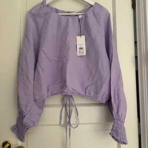 Ljuslila blus med knyt  - Supersöt ljuslila blus med långa ärmar och smockade muddar. Blusen har knytband både i midjan och bak, samt en öppen detalj i ryggen, perfekt för vår och sommar. 