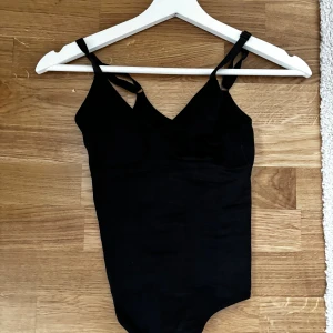 Svart ribbad body - Säljer en svart ribbad body med smala justerbara axelband och v-ringad hals. Toppen är tight och har en enkel, stilren design som passar perfekt till jeans eller kjol. Materialet är stretchigt och bekvämt.