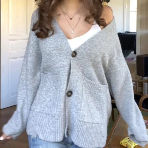 Grå kofta med stora knappar - Mysig ljusgrå kofta med v-ringning och tre stora, mönstrade knappar framtill. Koftan har två rejäla fickor och ribbade detaljer vid ärmslut och nederkant. Perfekt för lager-på-lager och chill dagar.
