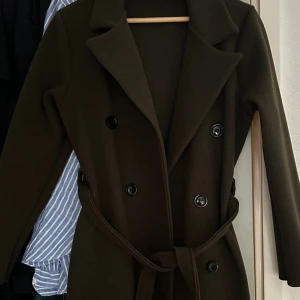 Olive  kappa med bälte och knappar - Stilren olive  kappa med dubbelknäppning och bälte i midjan. Perfekt för kyligare dagar.