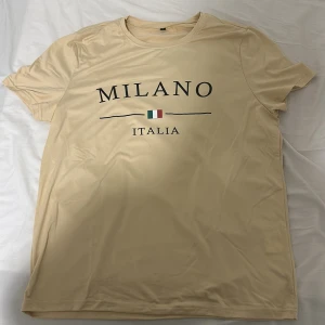 Beige Milano t-shirt med tryck - Beige t-shirt med svart text 'MILANO ITALIA' och en liten italiensk flagga på bröstet. Klassisk rund hals och korta ärmar. T-shirten har en clean look och är gjord i ett mjukt, slätt material som känns skönt mot huden.