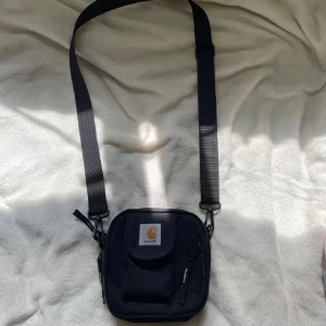 Svart axelväska från Carhartt - Säljer en svart axelväska från Carhartt med justerbar rem och flera fack med dragkedja. Väskan har en fyrkantig form och är tillverkad i slitstark polyester. Perfekt för att bära det viktigaste och har en tydlig Carhartt-logga framtill.